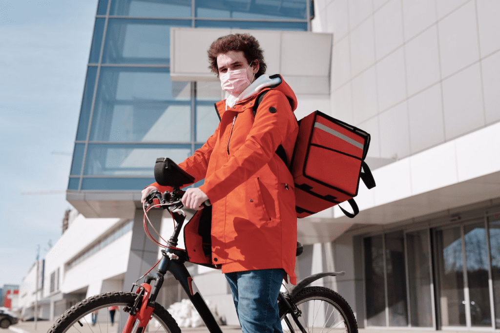Entregador de bike de máscara e mochila térmica. Foto: Pexels