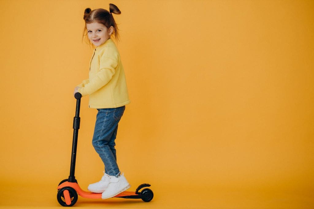 Imagem representando o melhor patinete infantil. Menina pequena, com 5 ou 6 anos de idade, pele clara, cabelos castanhos presos em dois coques, usando casaco amarelo e calça jeans, vista de perfil e de corpo inteiro, sorri com o rosto virado para a câmera, em cima de patinete infantil preto e laranja. O fundo é amarelo. Imagem: @senivpetro, Freepik.com.